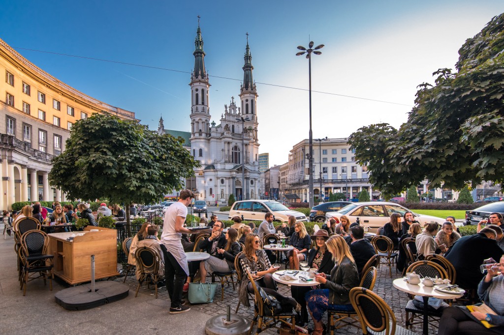 Plac Zbawiciela, na pierwszym planie ludzie siedzą przy stolikach w ogródku restauracyjnym, w tle kościół Najświętszego Zbawiciela, kamienice, samochody, dzień, lato.
