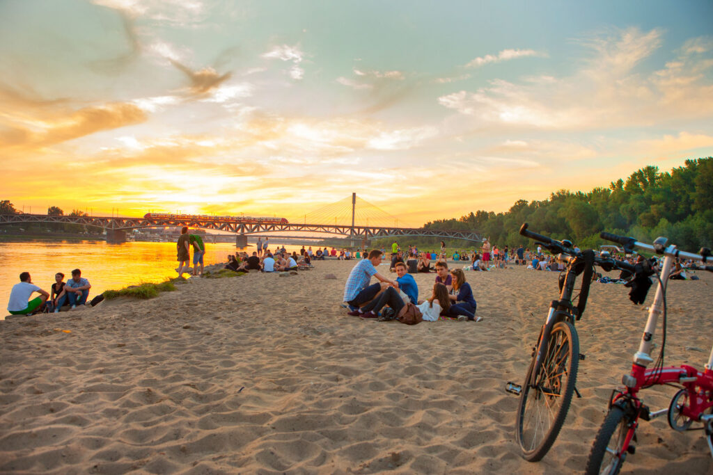 Grupa młodych ludzi siedzi na piaszczystej plaży na tle zachodzącego słońca. Na pierwszym planie zaparkowany rower.
