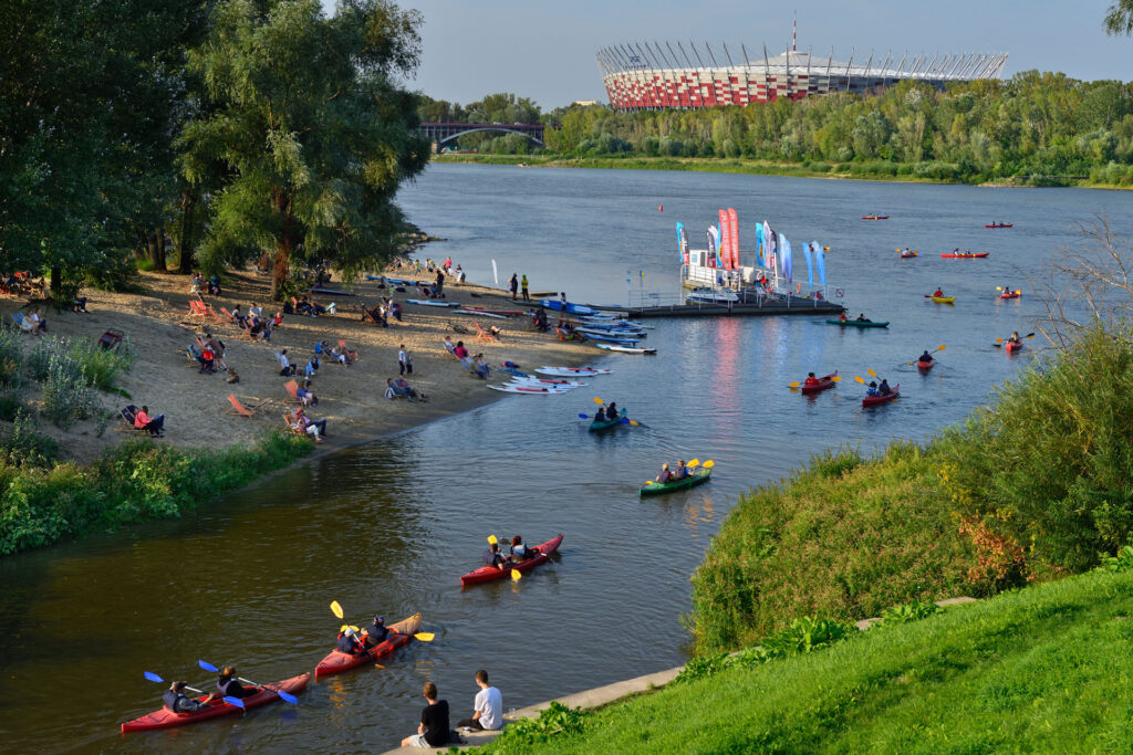 Spływ kajakowy po Wiśle. Na plaży ludzie wypoczywają w leżakach. W tle Stadion Narodowy i zieleń prawego brzegu Wisły.