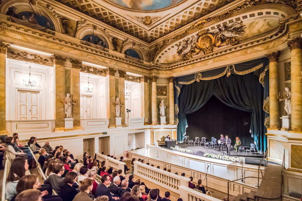 Barokowe wnętrze Teatru Królewskiego, widownia zapełniona, na scenie przemawiają artyści.