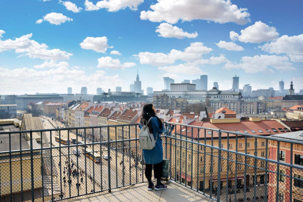 Kobieta ogląda panoramę Warszawy z wysoko położonego tarasu. Na pierwszym planie kamienice Krakowskiego Przedmieścia, w tle nowoczesne wieżowce.