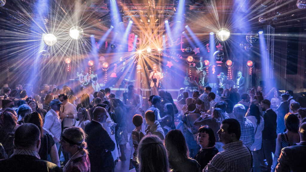 Koncert na sali ozdobionej dyskotekowymi kulami rozbłyskującymi światło. Na parkiecie ludzie w średnim wieku.