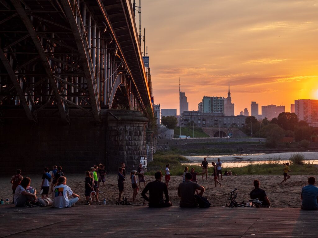 Zachód słońca na miejskiej plaży, po lewej stronie most, na pierwszym planie siedzą ludzie .