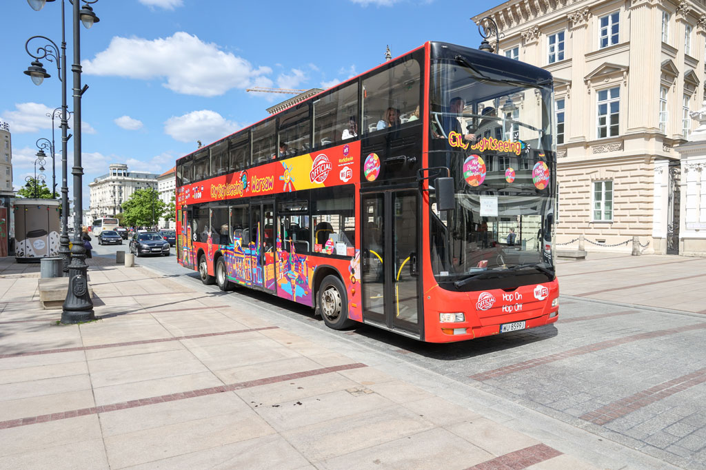Dwupoziomowy, czerwony autobus turystyczny jedzie ulicą Krakowskie Przedmieście, za nim jadą samochody, w tle kamienice z kremową elewacją, słoneczny dzień, lato.