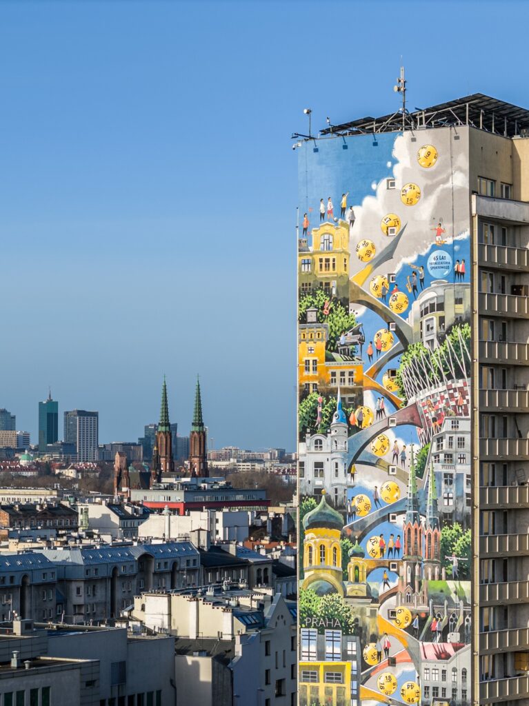 Barwny mural przedstawiający reprezentatywne obiekty Pragi, umieszczony na całej ścianie wieżowca, w tle panorama warszawskiej Pragi