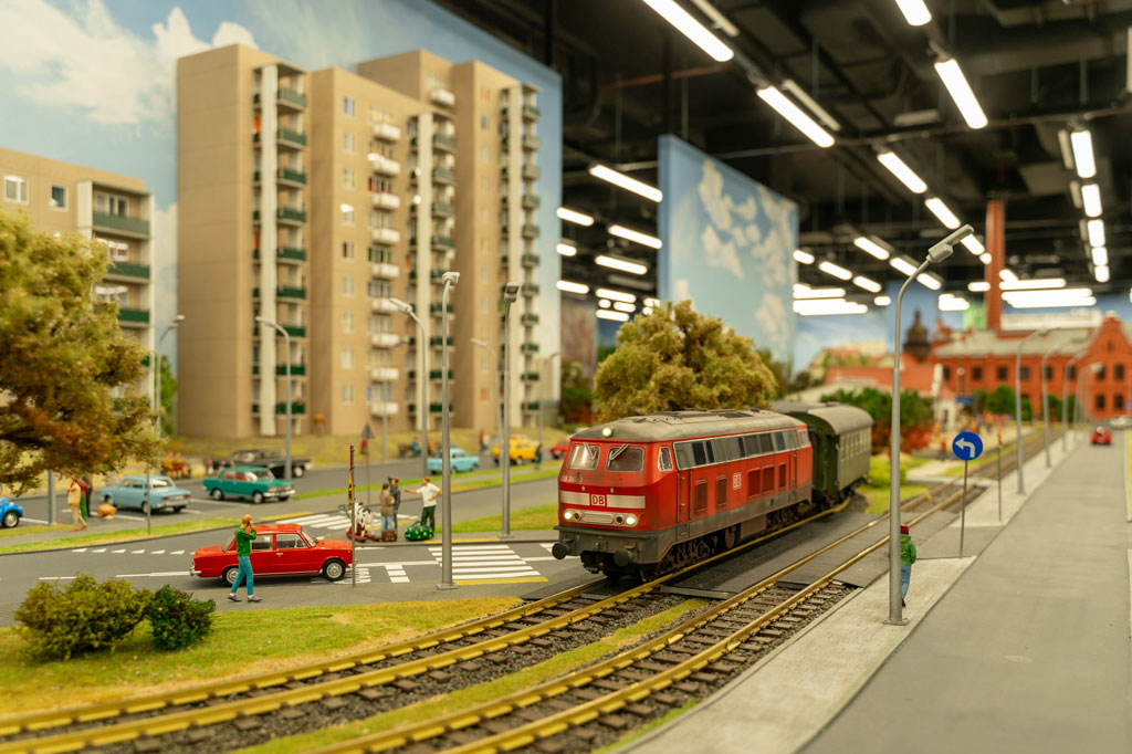 Kolejkowo, makieta miasta, na pierwszym planie jedzie pociąg, po lewej stronie wysokie bloki, dalej ceglane zabudowania fabryczne, obok stoją miniaturowe samochodziki, dookoła małe postaci ludzi.
