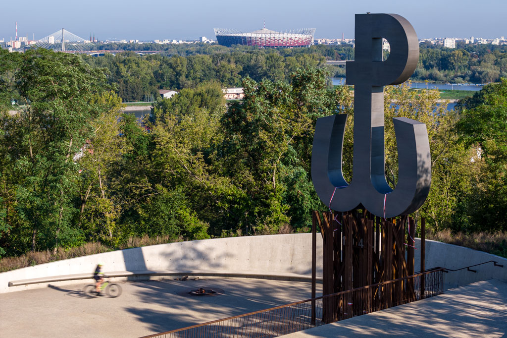 Betonowy taras na szczycie kopca Powstania Warszawskiego, duży znak Polski Walczącej umieszczony na metalowych prętach, dookoła drzewa, w tle stadion PGE Narodowy, most świętokrzyski, budynki, słoneczny dzień, lato.