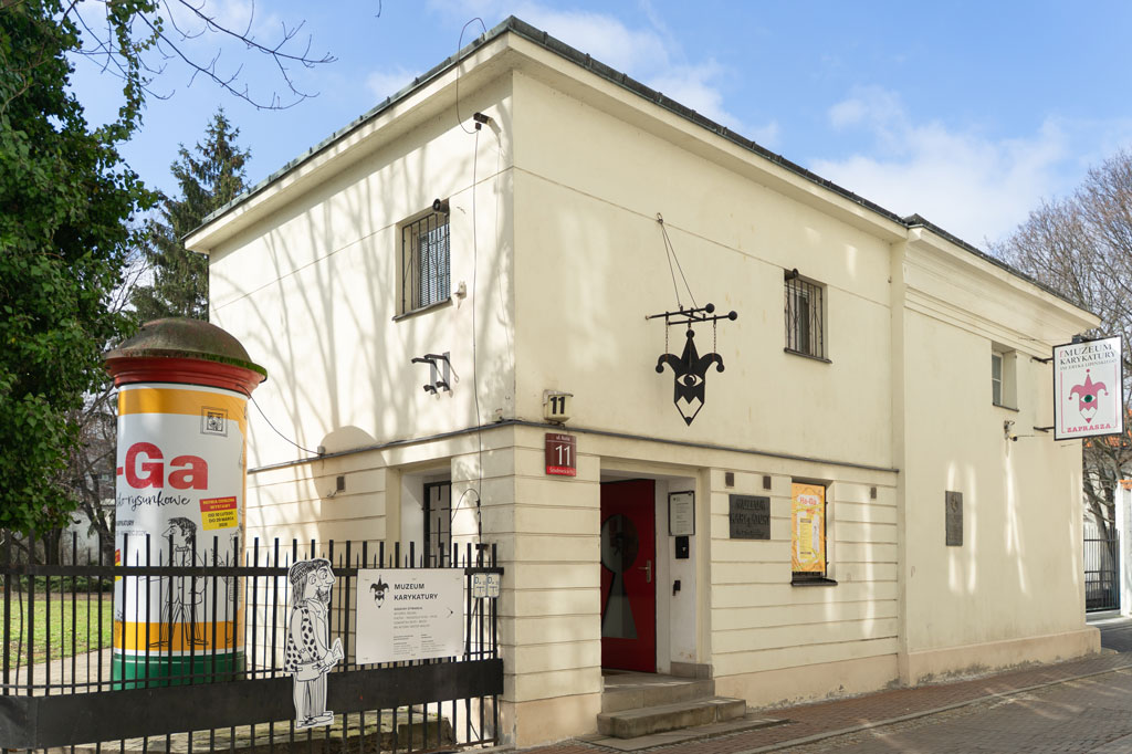Widok na dwupiętrowy, jasny budynek Muzeum Karykatury. Nad wejściem wisi szyld w kształcie głowy błazna zakończonej ostrzem ołówka. Przy wejściu znajduje się czerwona tabliczka z numerem 11 oraz słup ogłoszeniowy z plakatem wystawy „Ha-Ga”.