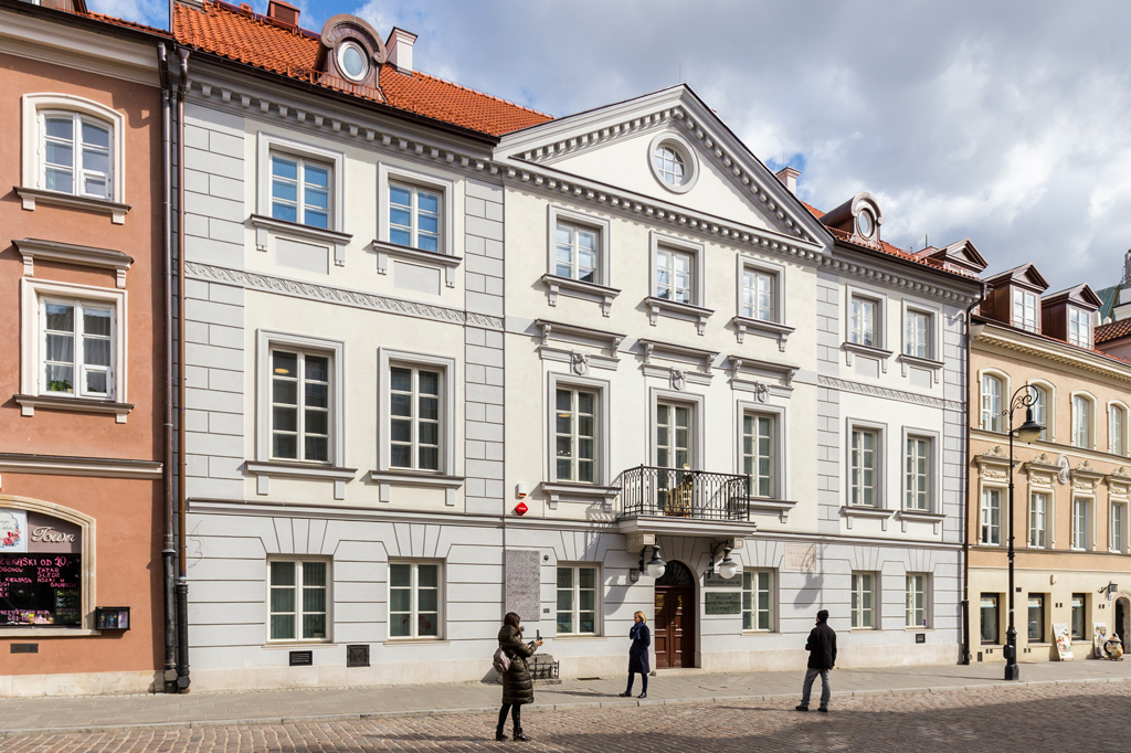Dwupiętrowa, zabytkowa, biała kamienica z balkonem na pierwszym piętrze. Przed nią stoi kilka osób.
