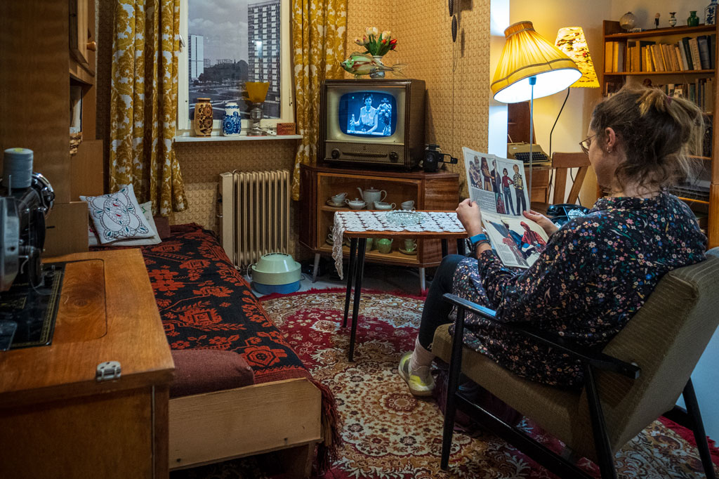 Pomieszczenie zaaranżowane na pokój z wystrojem z lat 70./80. XX wieku, meblościanka, na szafce w rogu stoi telewizor, na nim wazon ze sztucznymi kwiatami. Na fotelu siedzi kobieta, przegląda kolorowe czasopismo.