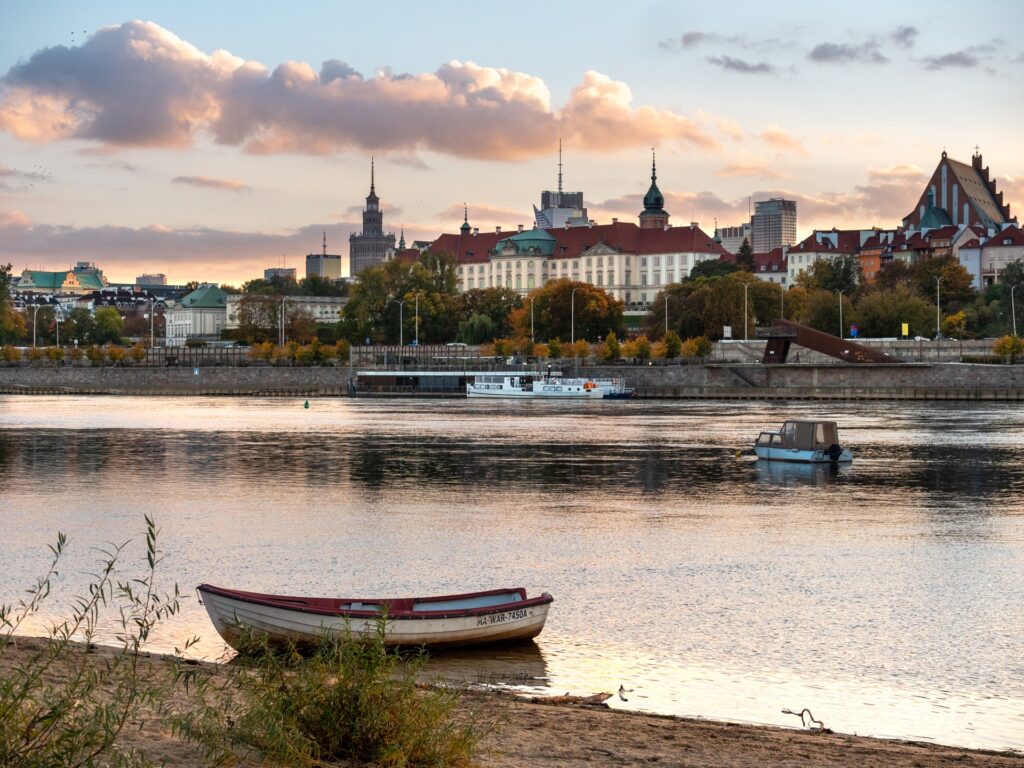 Łódka zacumowana przy plaży nad Wisłą, w tle Stare Miasto o zachodzie słońca.