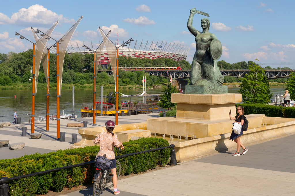Pomnik Syrenki nad Wisłą, spacerowicze, rowerzyści, w tle Wisła, Stadion PGE Narodowy, po moście średnicowym jedzie pociąg, zieleń drzew i krzewów, słoneczny dzień, lato.