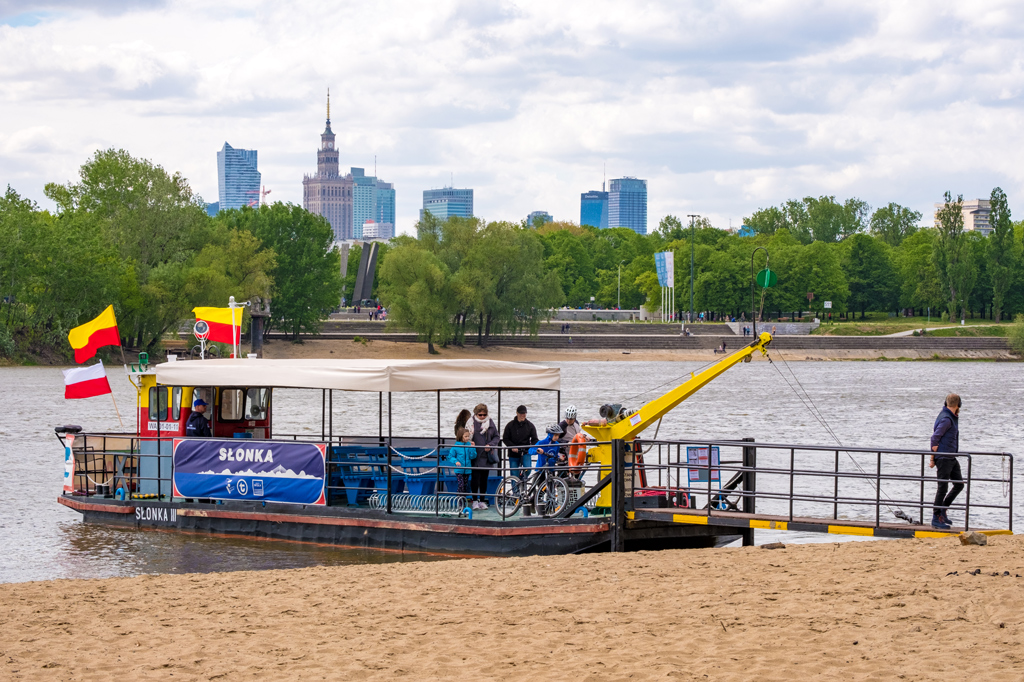Prom Słonka zacumowany przy plaży, w tle Pomnik Chwała Saperom, panorama centrum Warszawy, drzewa, zieleń, dzień, lato.