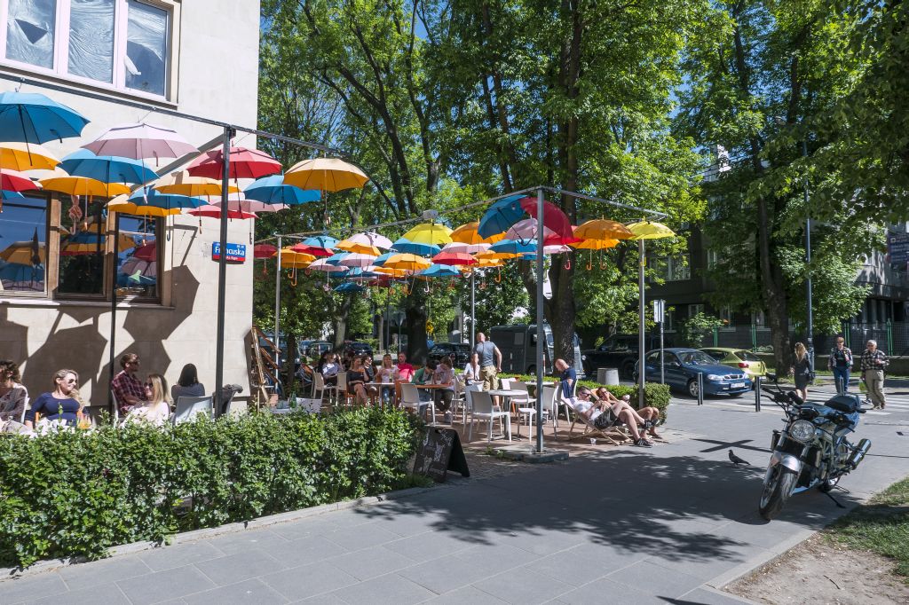 Ulica Francuska na Saskiej Kępie, ludzie siedzą przy stolikach i na leżakach w ogródku restauracyjnym, nad nimi wiszą kolorowe parasole, drzewa, zieleń, słoneczny dzień, lato.