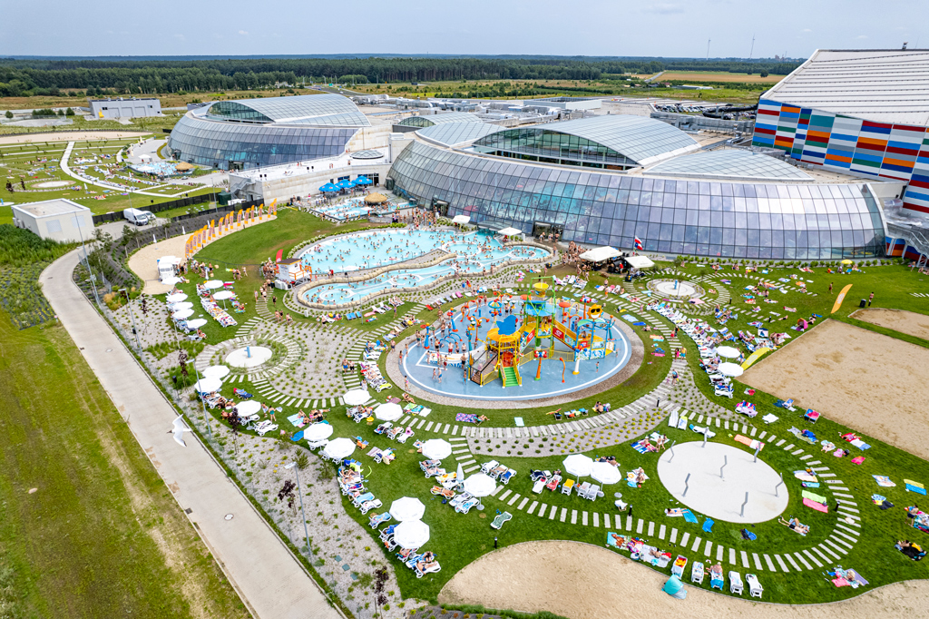 Widok z lotu ptaka na park wodny Suntago, ludzie pływają w basenach odkrytych, inni odpoczywają dookoła na leżakach i na trawie, dzieci bawią się na wodnym placu zabaw, obok baseny kryte pod szklanymi kopułami, dzień, lato.