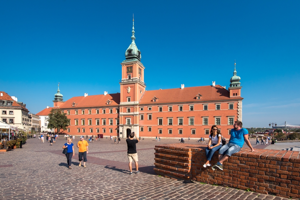 Podłużna, kilkupiętrowa, budowla ceglanych barwach z jedną większą wieżą pośrodku i dwoma mniejszymi wieżyczkami na rogach. Na brukowanym placu przed budynkiem spacerują ludzie. Na pierwszym planie na ceglanym murku siedzi młoda kobieta i mężczyzna. Nad budynkiem widać bezchmurne, niebieskie niebo.
